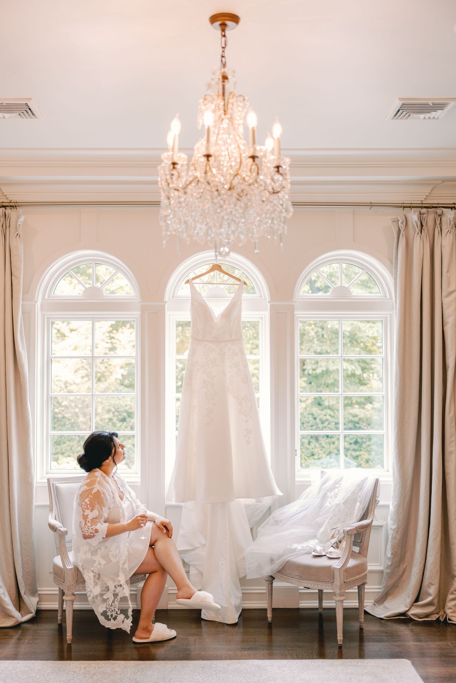 Bride in lace robe seated in a bright bridal suite with wedding gown hanging in the window.