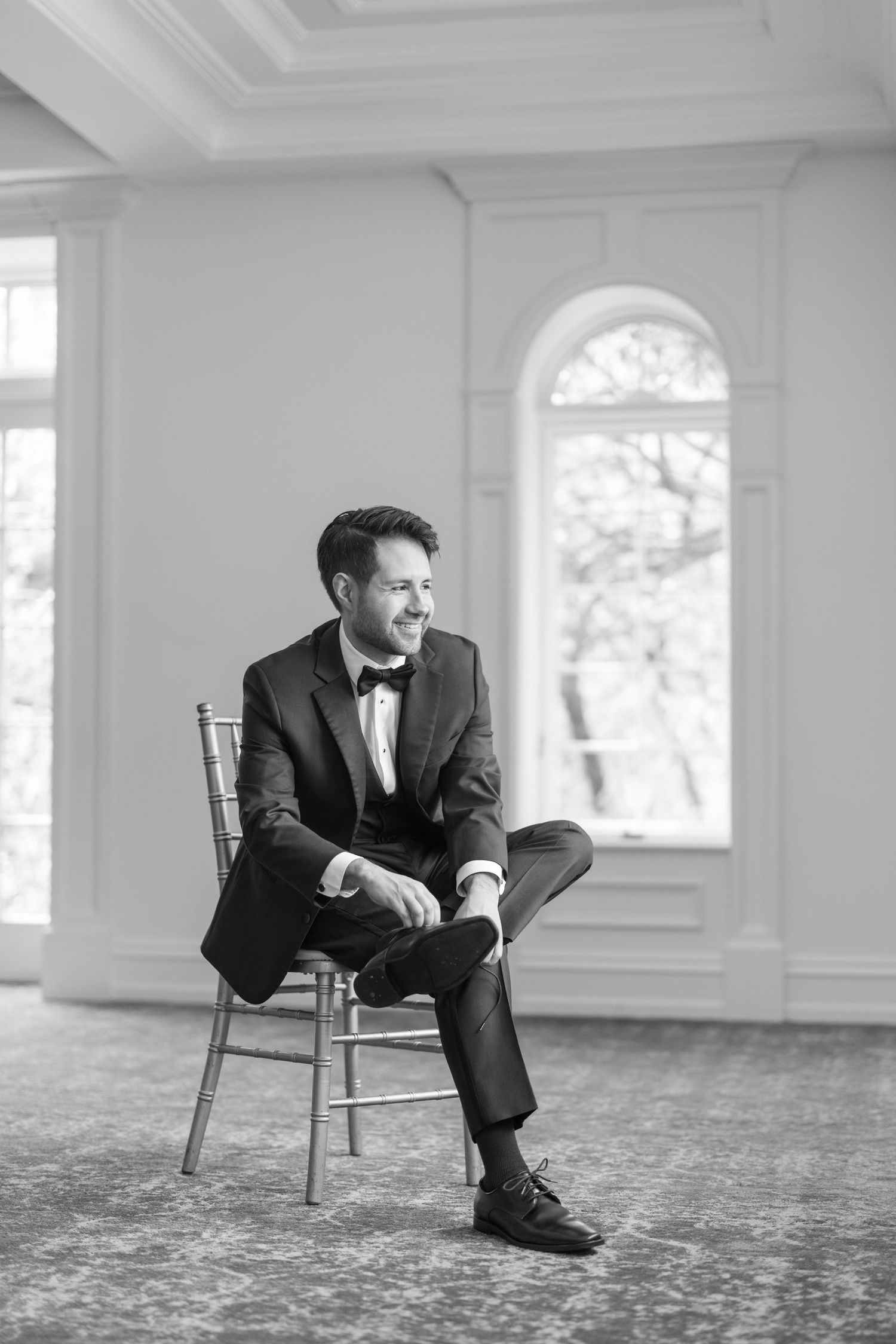 Dapper groom in black tuxedo and bow tie sits relaxed on chair in elegant ballroom.