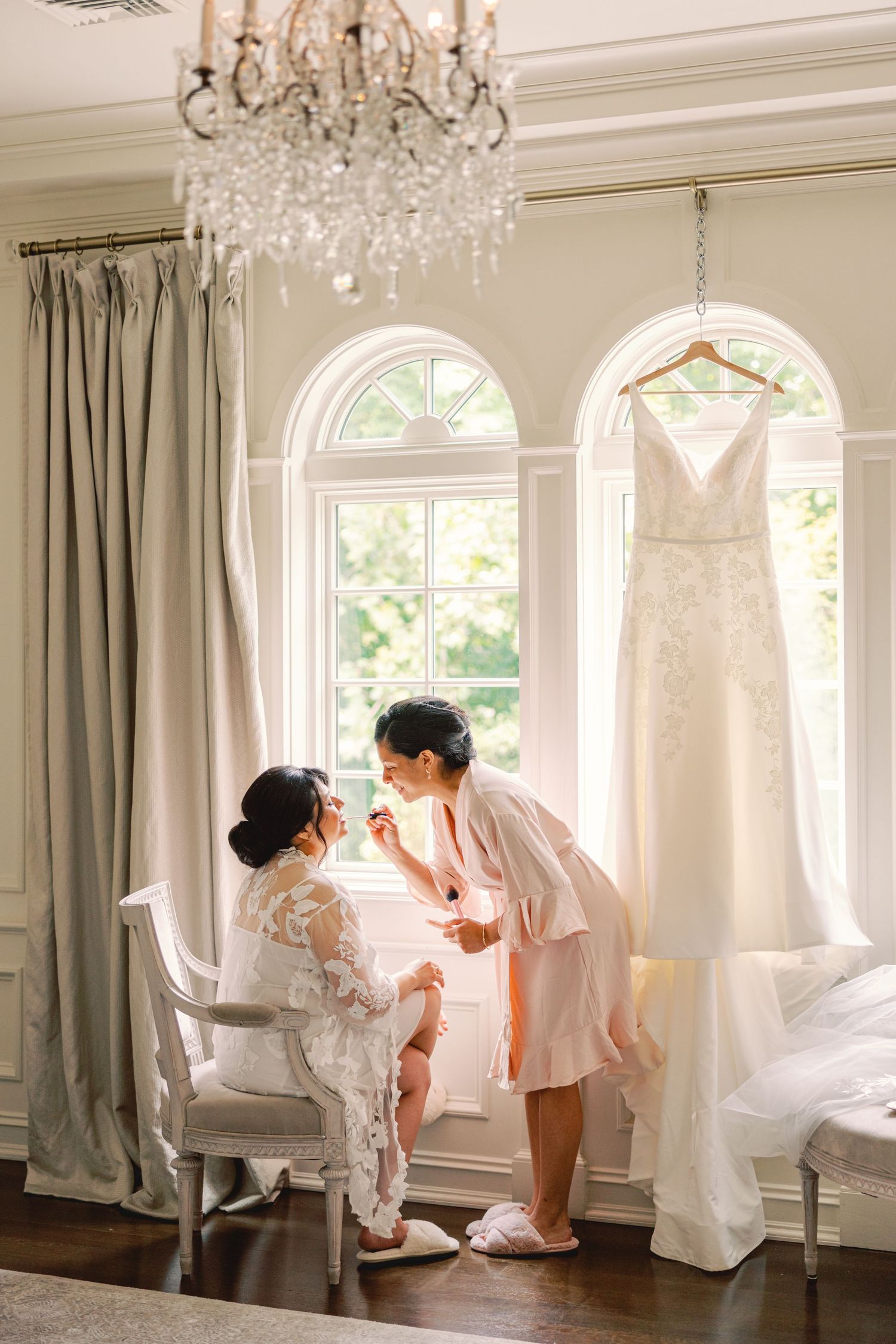 Bride and bridesmaid sharing a moment in a bright bridal suite with wedding dress hanging nearby.