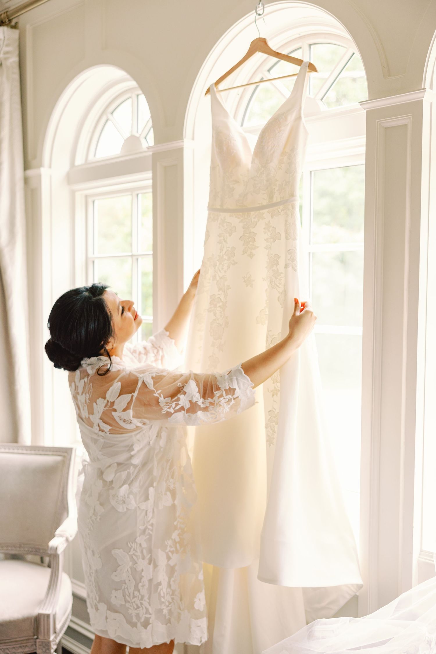 Bride in lace robe reaching up to touch her wedding gown hanging in a sunlit bridal suite.