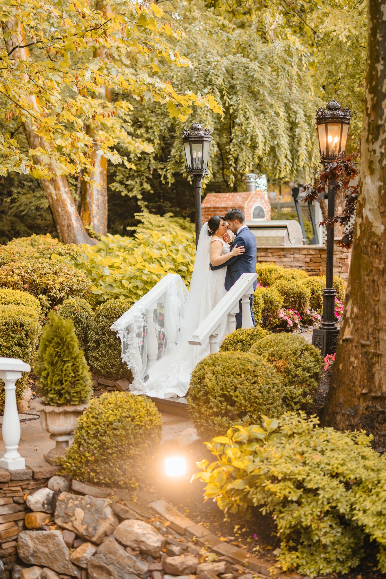Bride and groom share a romantic embrace in a lush garden with golden autumn foliage and vintage lanterns.
