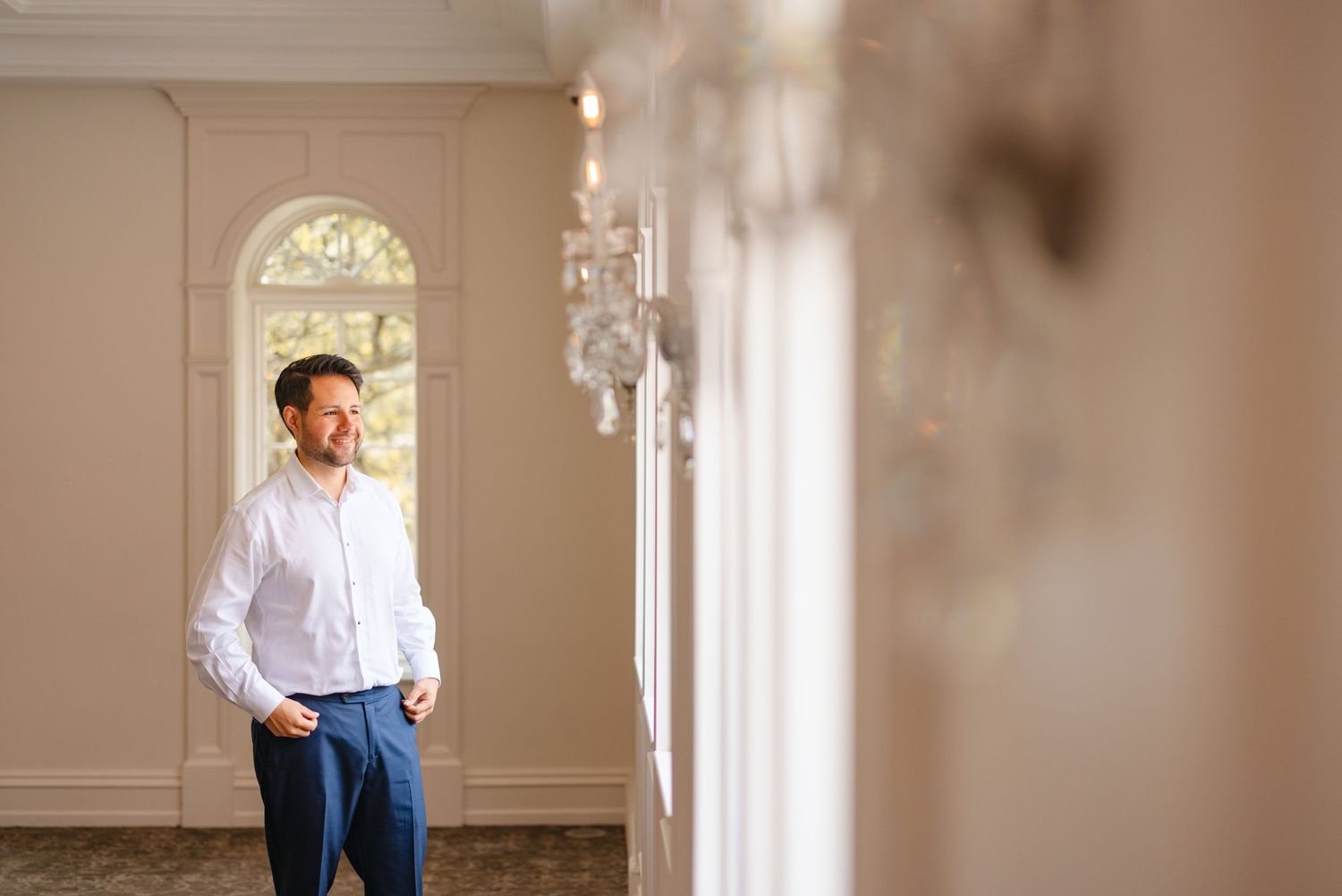 Groom in white shirt and blue trousers stands relaxed in elegant chandelier-lit wedding venue.