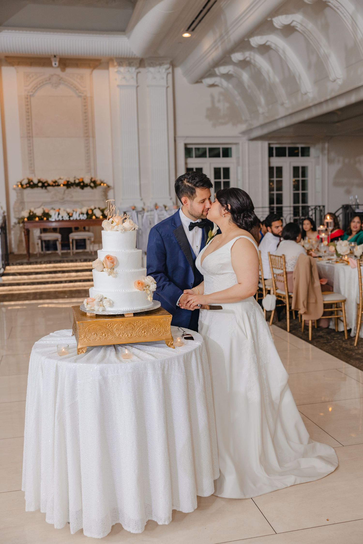 Bride and groom sharing a kiss while cutting their elegant four-tier white wedding cake at reception.