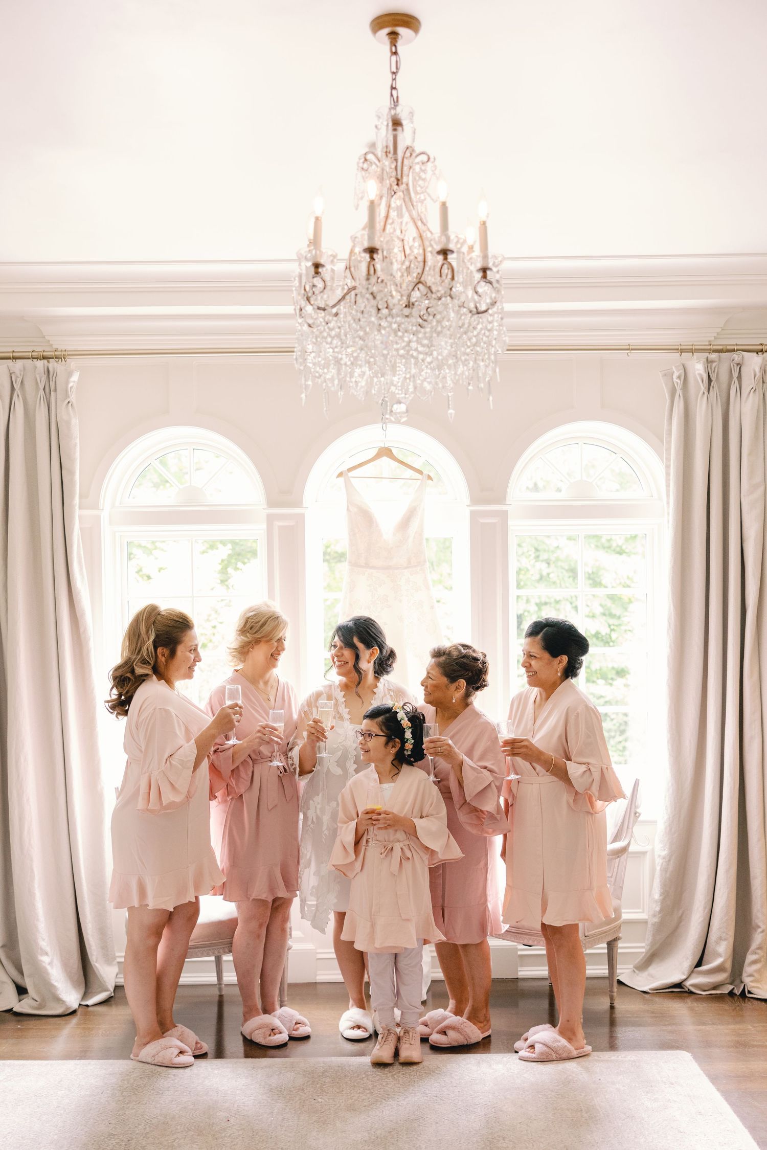 Bride and bridesmaids in matching pink robes toasting with champagne beneath a crystal chandelier.