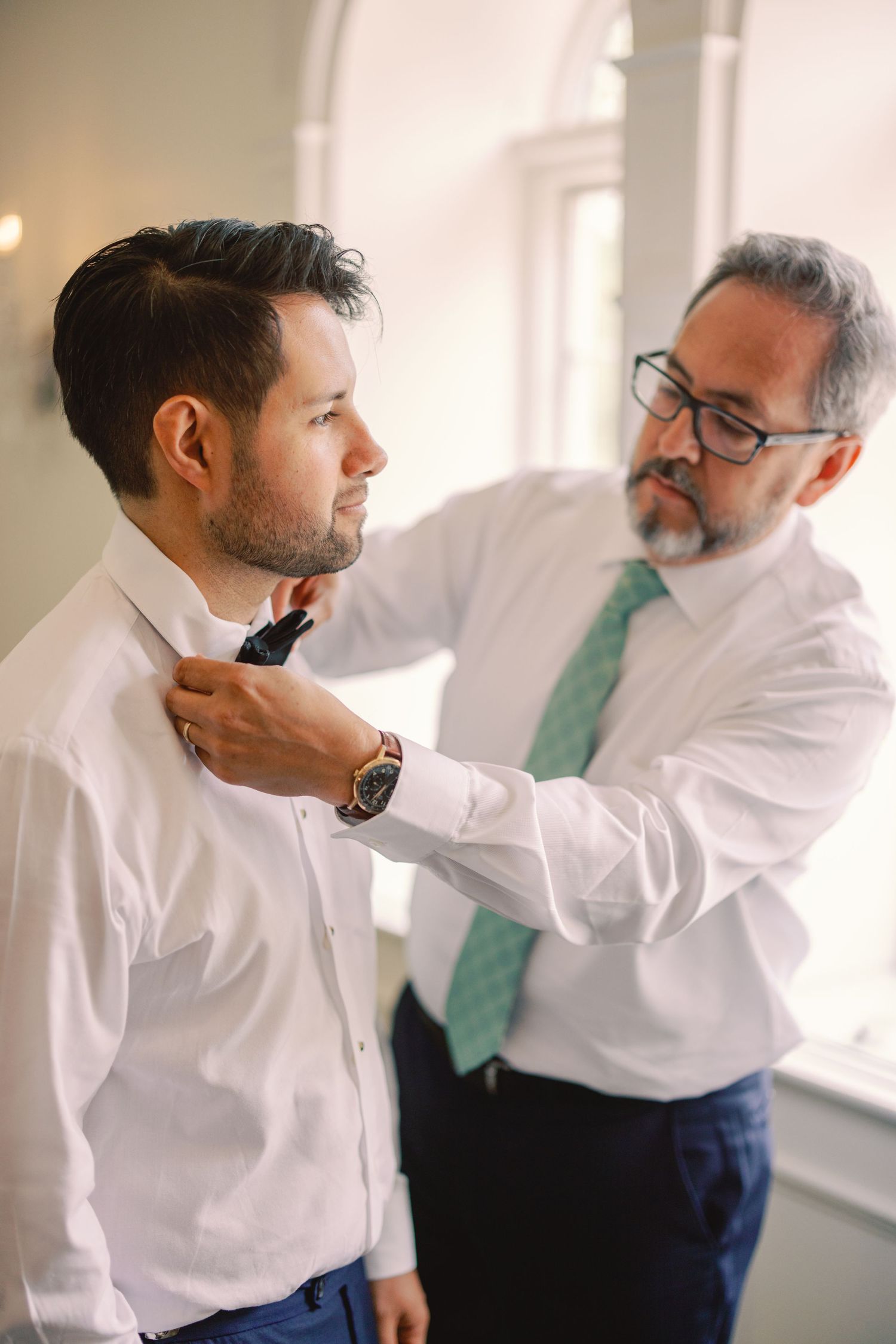 Father adjusts grooms bow tie as groom prepares for wedding day in white dress shirt.