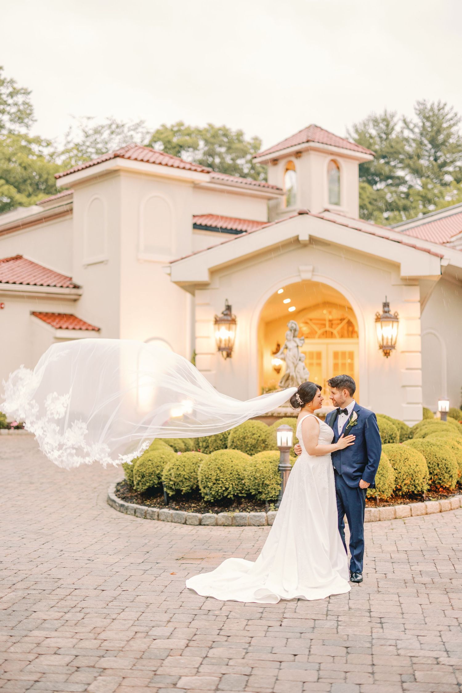 Bride and groom kissing as wedding veil flows dramatically in the wind outside an elegant venue entrance.