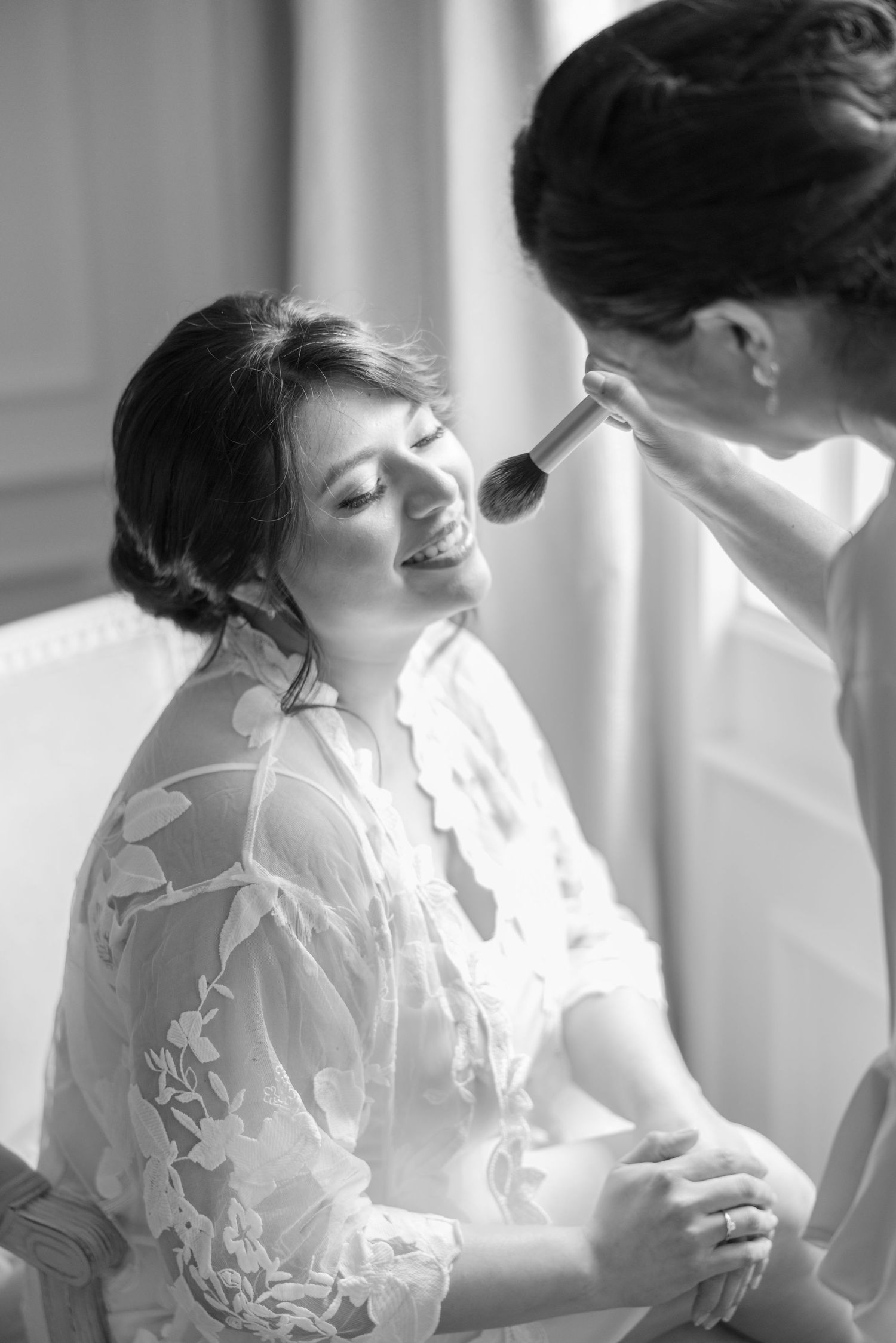Black and white photo of smiling bride getting makeup applied by artist on wedding morning.