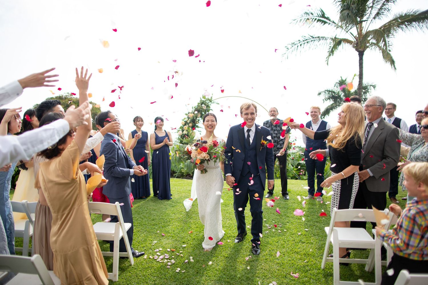 Wedding guests throw flower petals at an outdoor ceremony with palm trees and chairs.