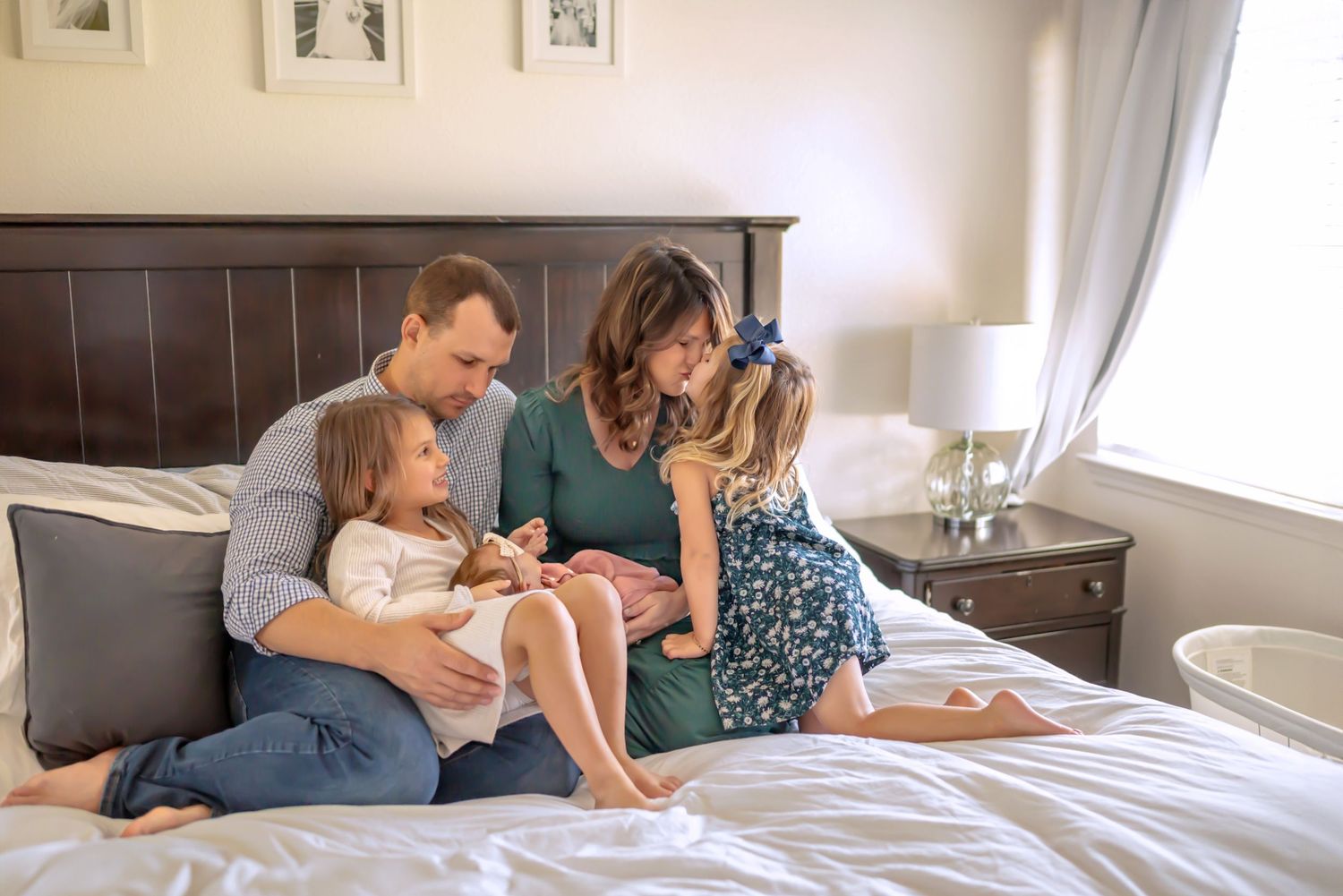 Family on bed together. Dad holds oldest. Mom gets kissed on nose by middle child and the newborn is snuggled between them. Doughman Photo