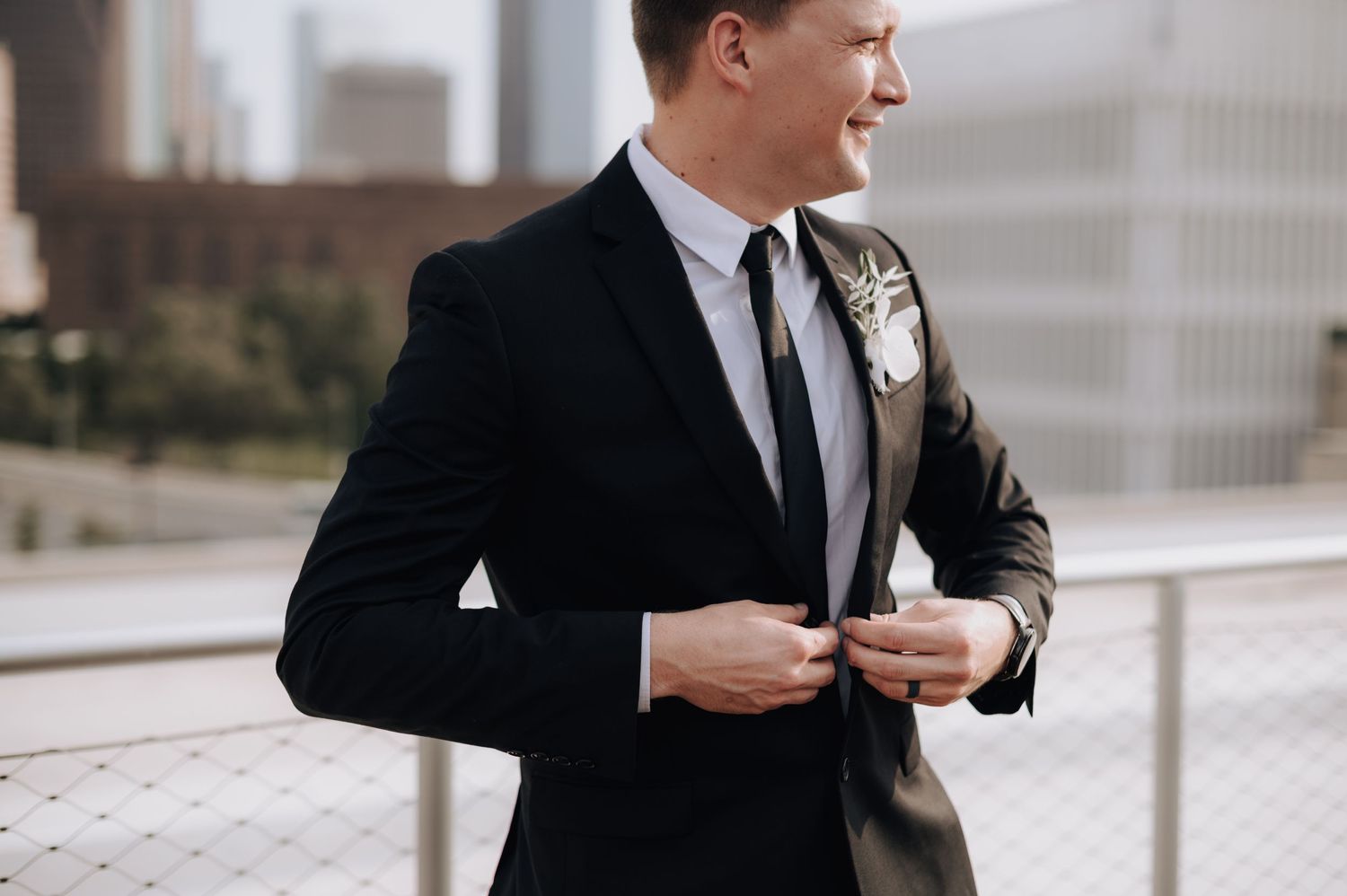 Groom in classic black suit adjusts his jacket while standing outdoors.