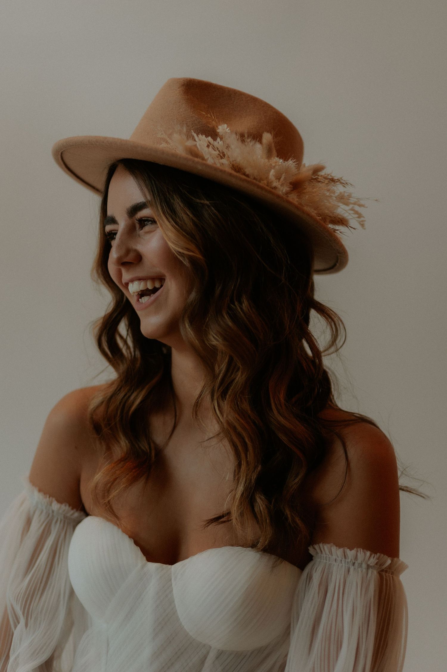 A romantic bridal portrait showcasing an off-shoulder wedding dress and brown felt hat with feather details.