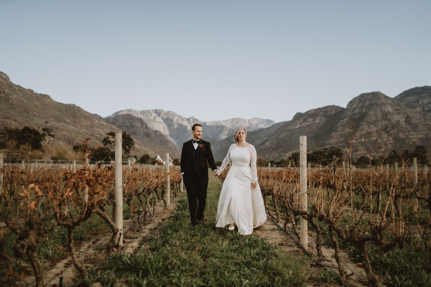 A couple walks through a scenic vineyard with mountains in the background during sunset.