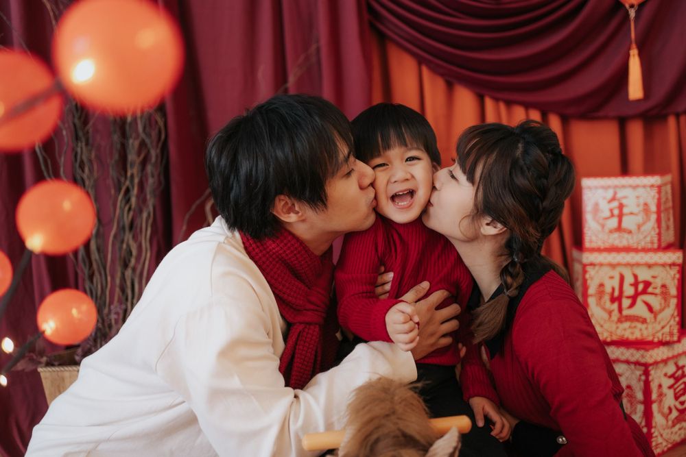 Family sharing tender moment with kisses against festive red Chinese New Year backdrop.