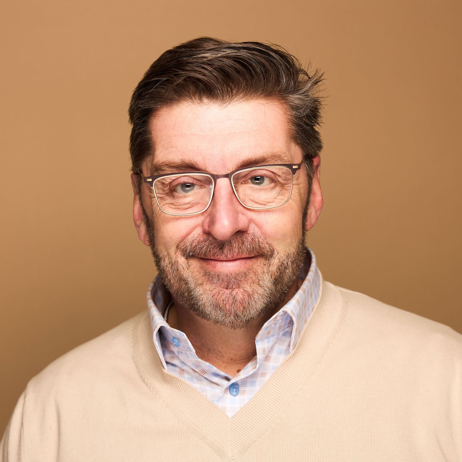 Professional headshot of person in glasses and beige sweater smiling against brown background.