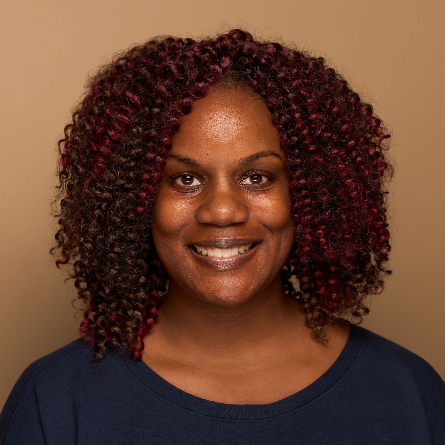 Portrait of person with vibrant burgundy curly hair wearing navy top against brown backdrop.