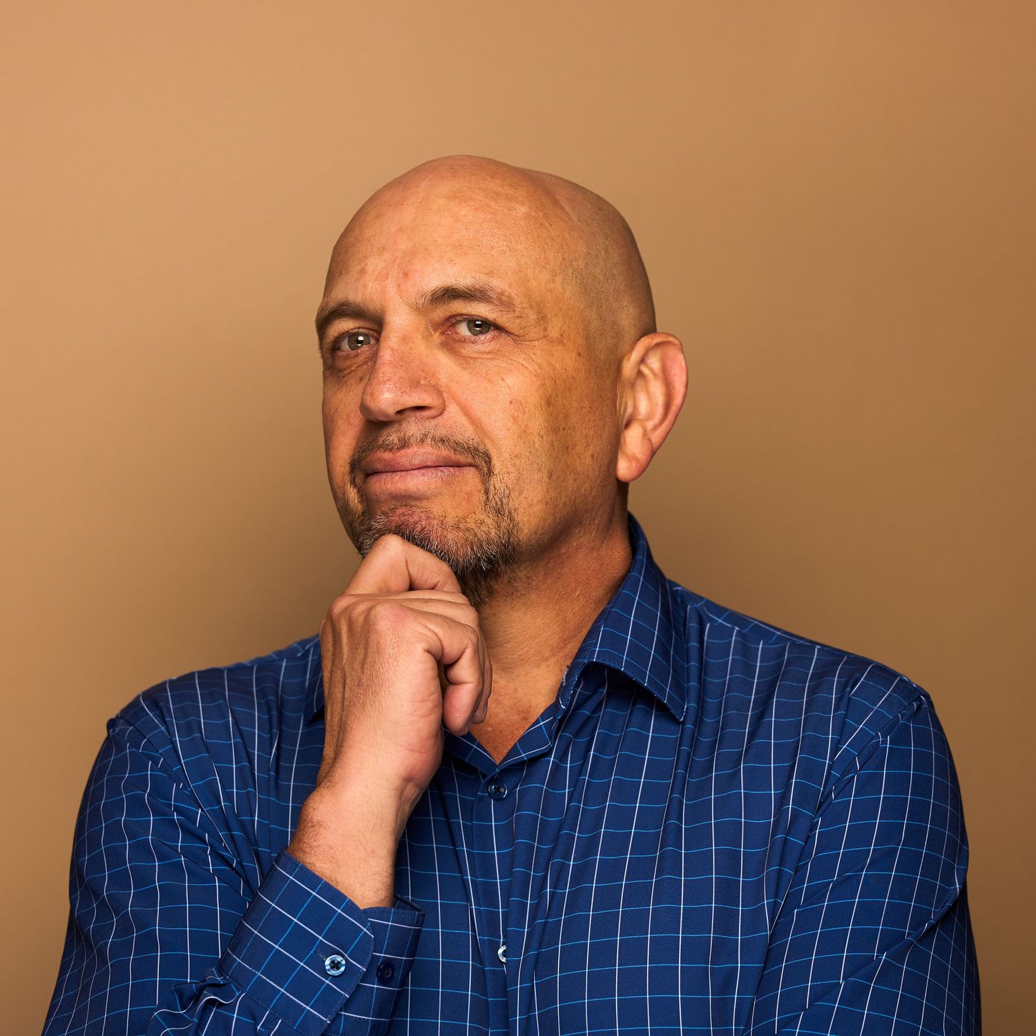Professional headshot of person in blue checkered shirt with hand on chin against brown background.