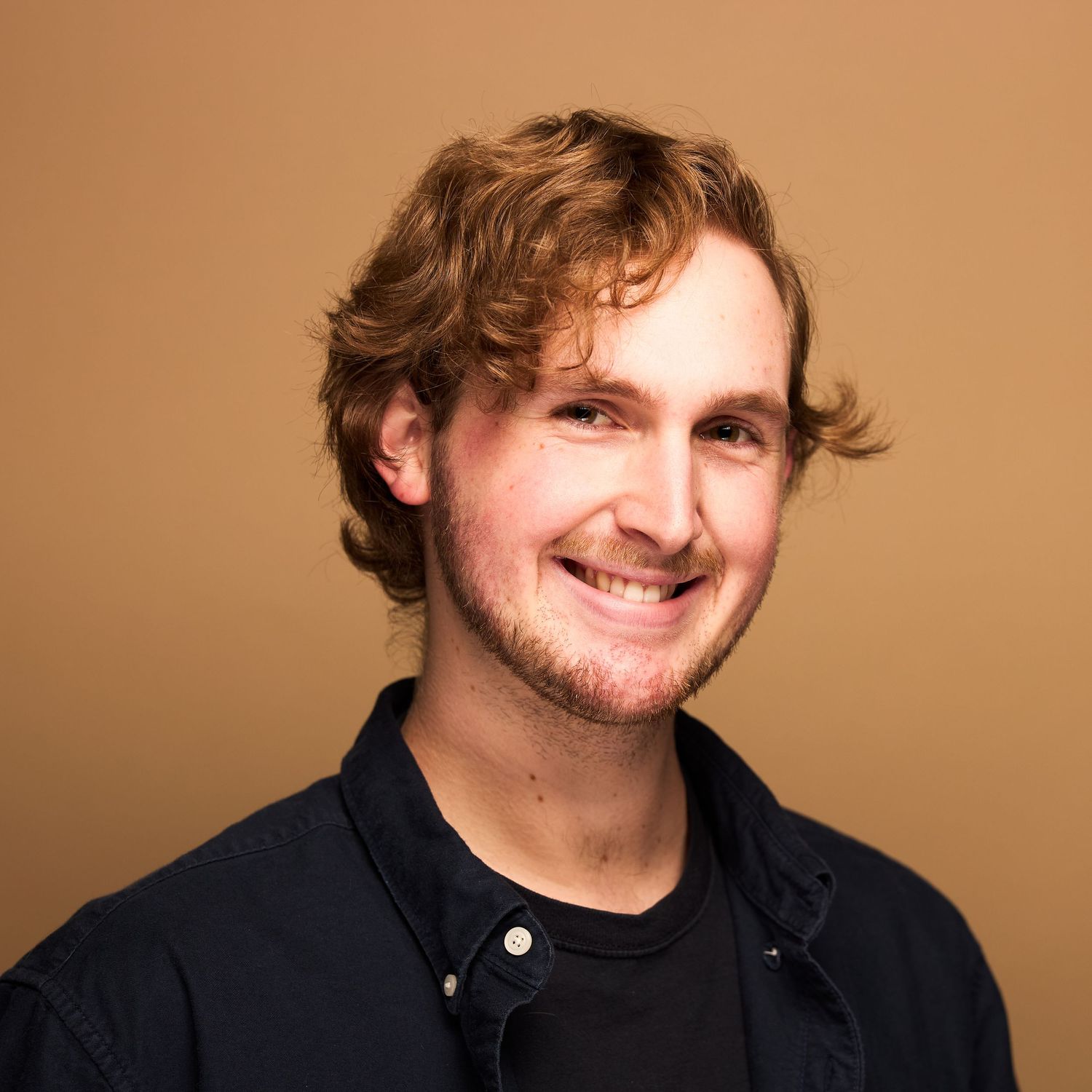 Portrait of smiling person with wavy hair wearing dark jacket against warm brown backdrop.