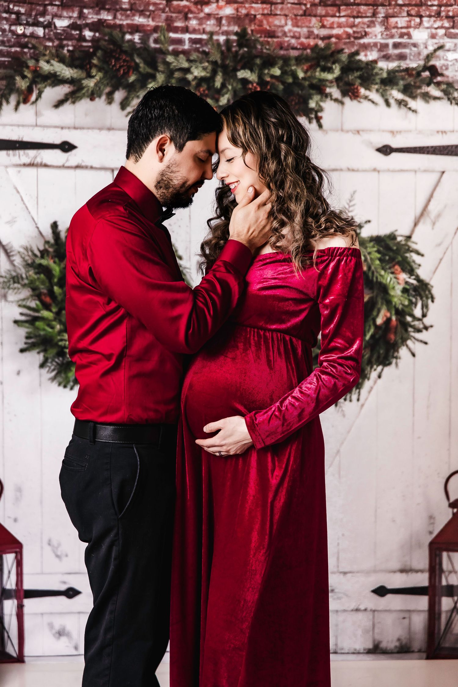 A romantic maternity photo of a couple in red attire embracing against a white barn door with evergreen garland.