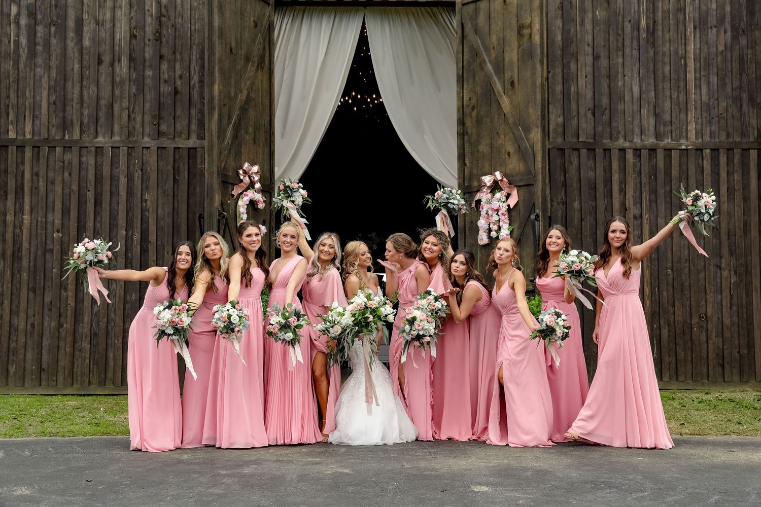 bridal party wearing pink bridesmaid dresses pose together in front of rustic barn entrance.
