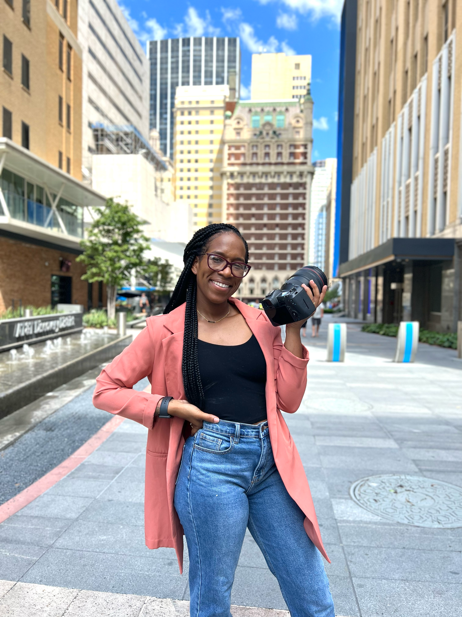 A photographer in a coral blazer poses with her camera in an urban setting with tall buildings.
