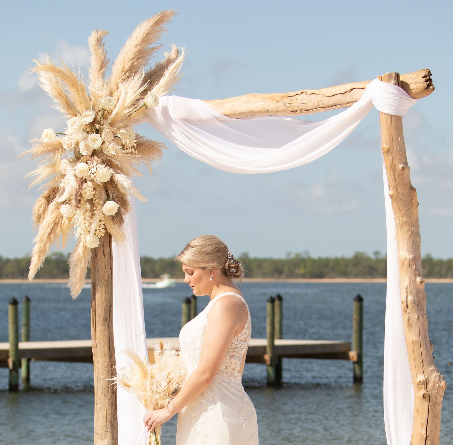 Beach wedding arch decorated with pampas grass and white fabric overlooking a waterfront dock.
