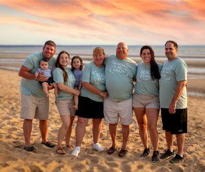 Family group wearing matching blue shirts poses together on sandy beach at sunset.