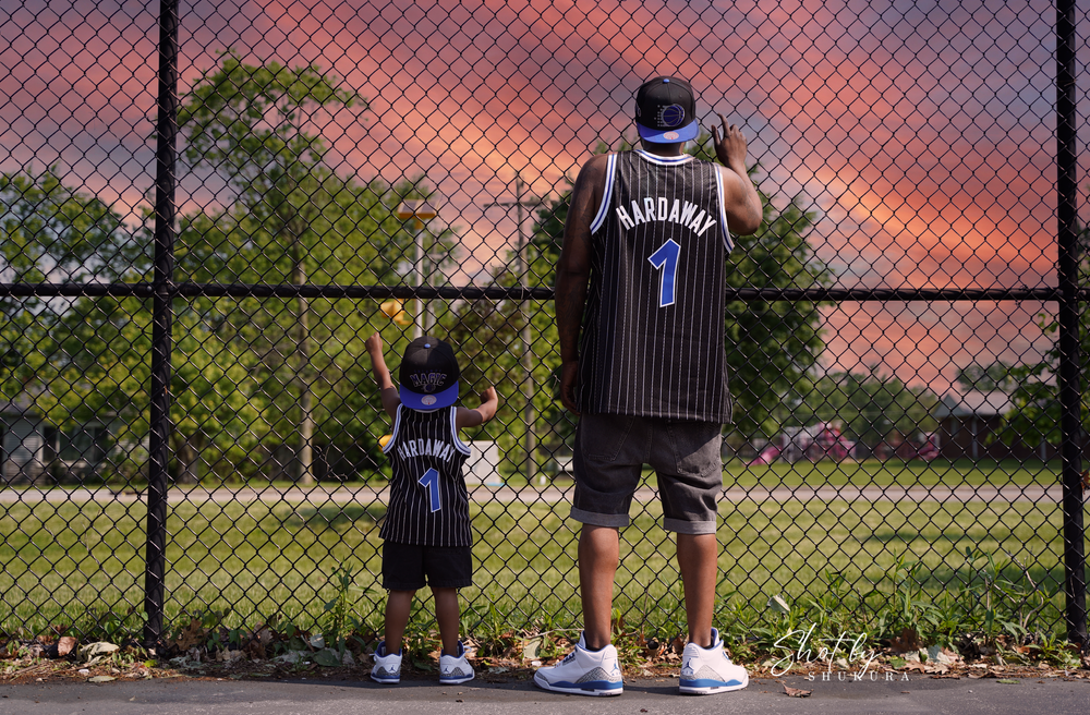 Two people in matching Orlando basketball jerseys stand looking at a chain link fence during sunset.