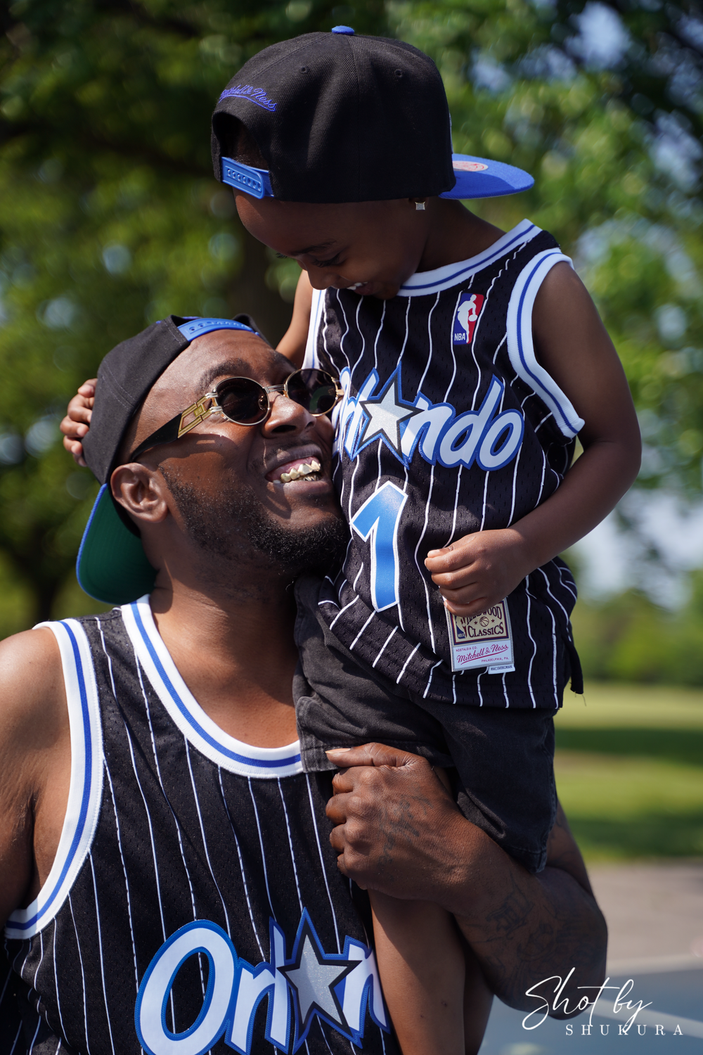 A father-child moment wearing matching Orlando Magic basketball jerseys outdoors on a sunny day.