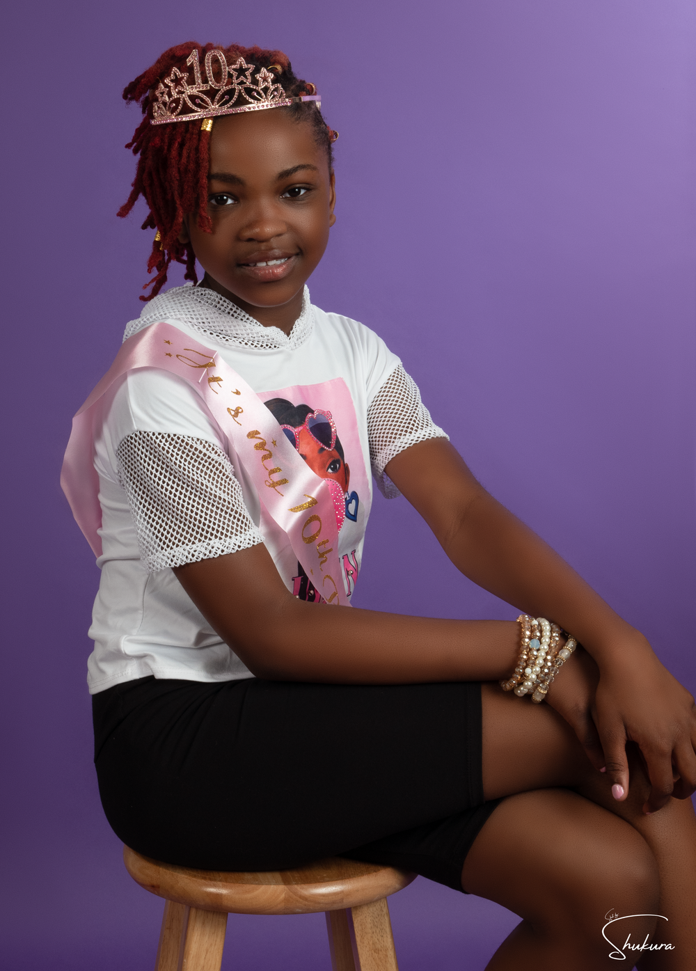 Youth portrait against purple background wearing white mesh top and black shorts with crown accessory.