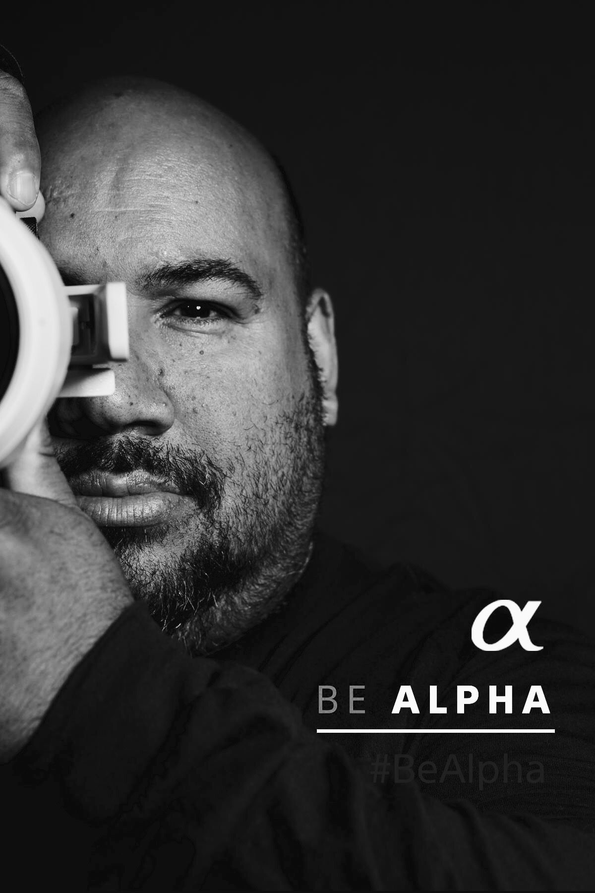 Black and white portrait of a photographer holding a camera lens in front of his eye with the text Be Alpha.