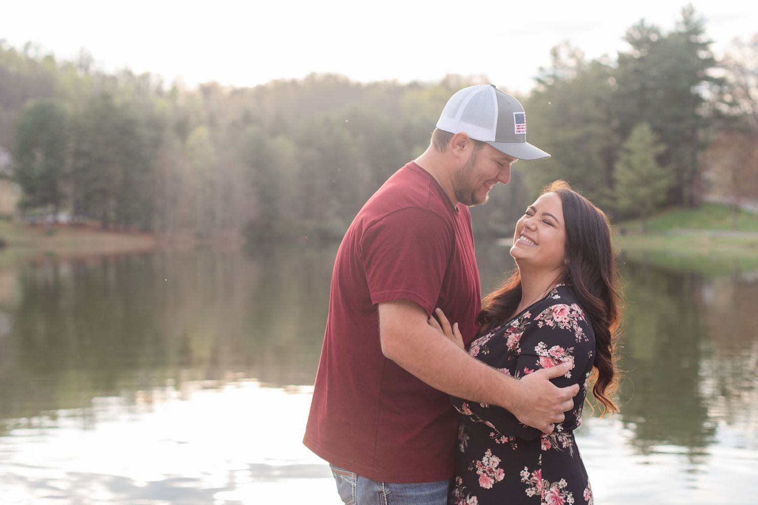Couple shares intimate moment by lakeside with pine trees in background during golden hour.
