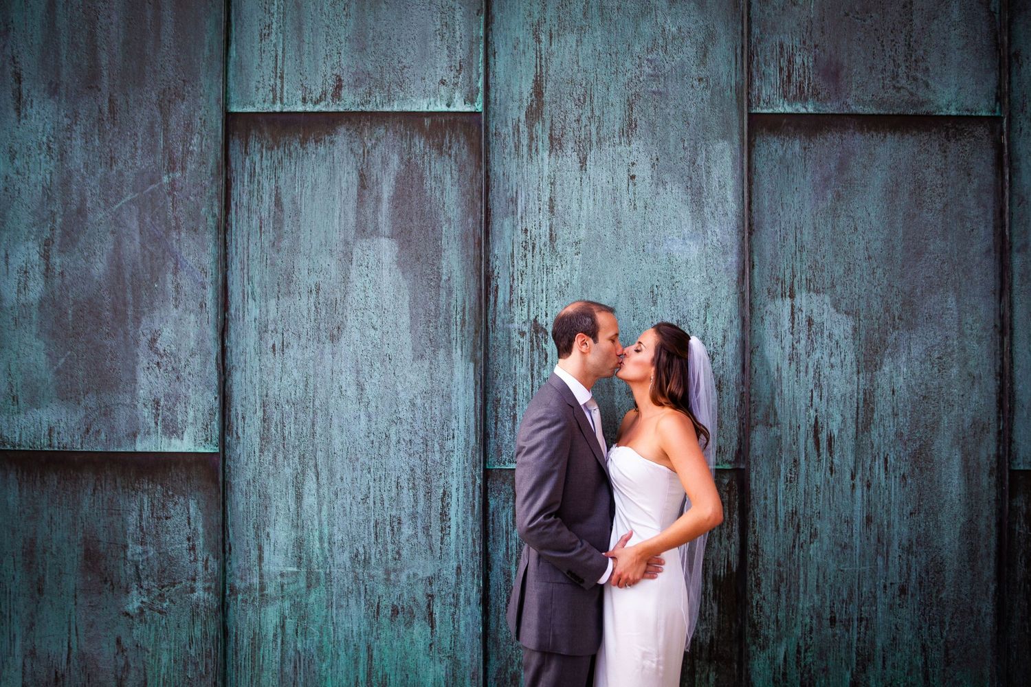 A couple poses against a weathered blue-green metal wall in formal wedding attire.