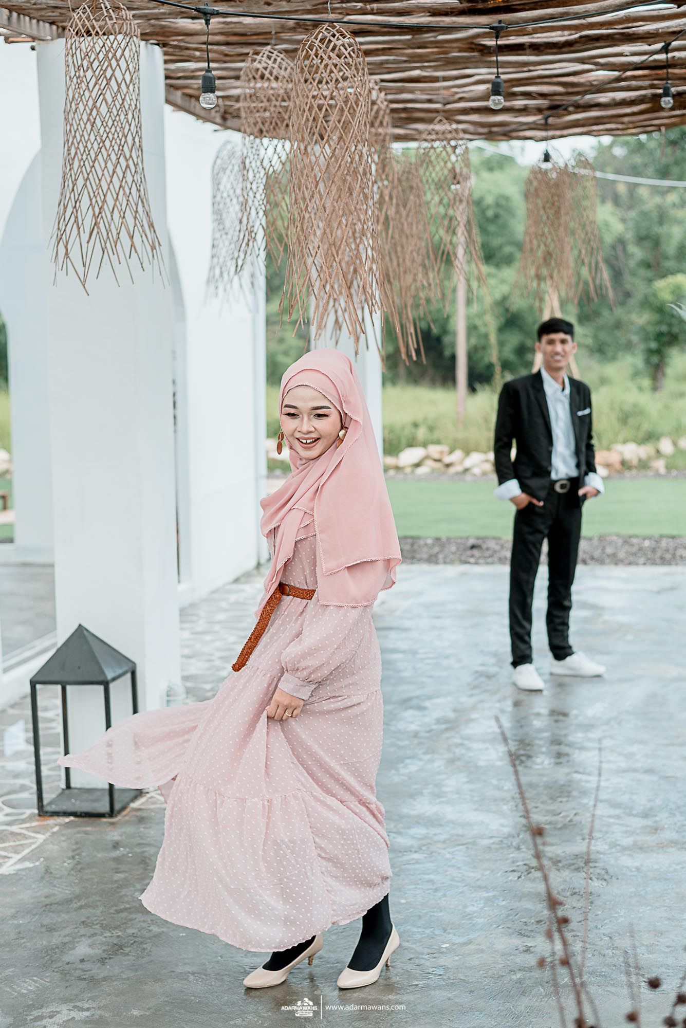 A couple poses under woven pendant lights with a woman in pink dress twirling and a man in black suit watching.