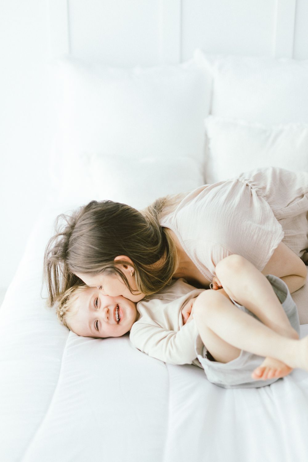 Parent and child sharing a joyful moment while laying on white bedding in bright natural light.