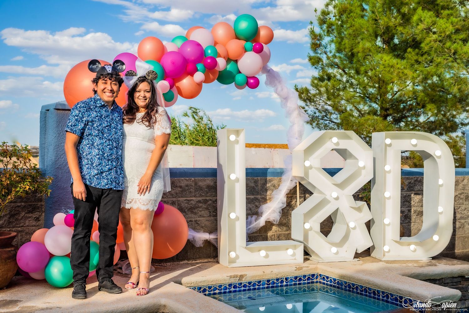 A poolside engagement photo with colorful balloon arch and large light-up initials in the background.