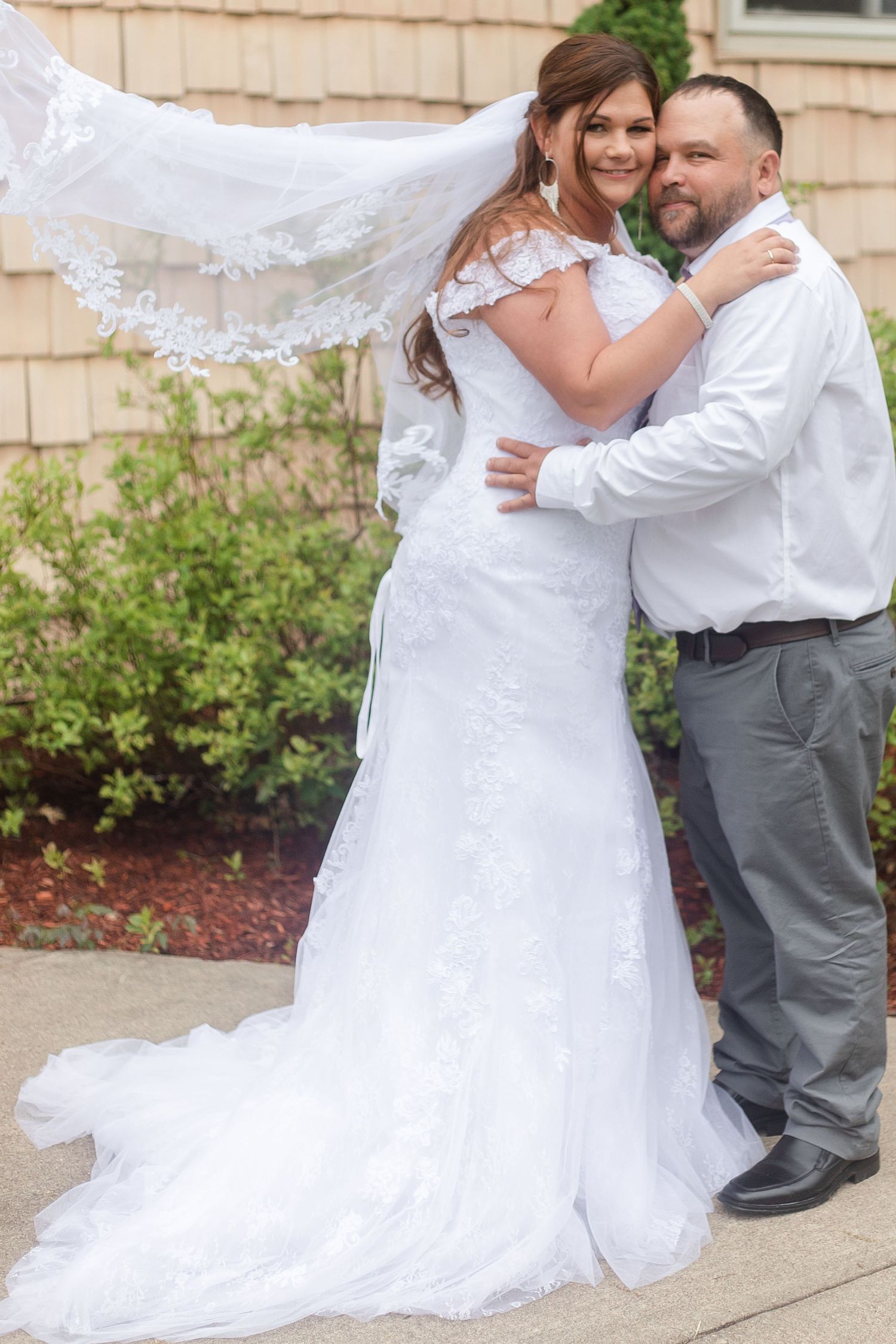 Wedding couple sharing intimate moments in white dress and formal attire against beige building.