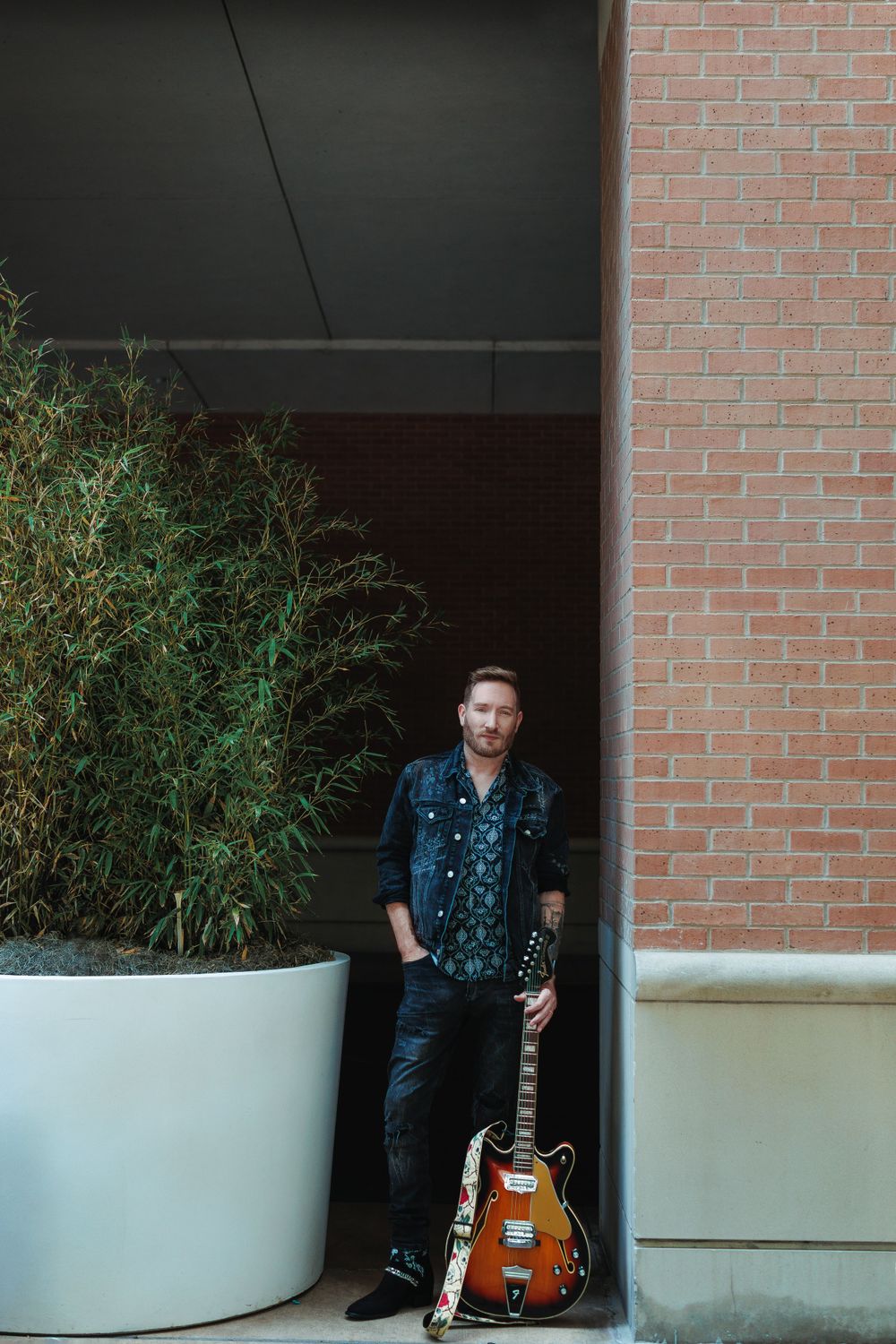 Musician posing with guitar against brick wall and modern planters.