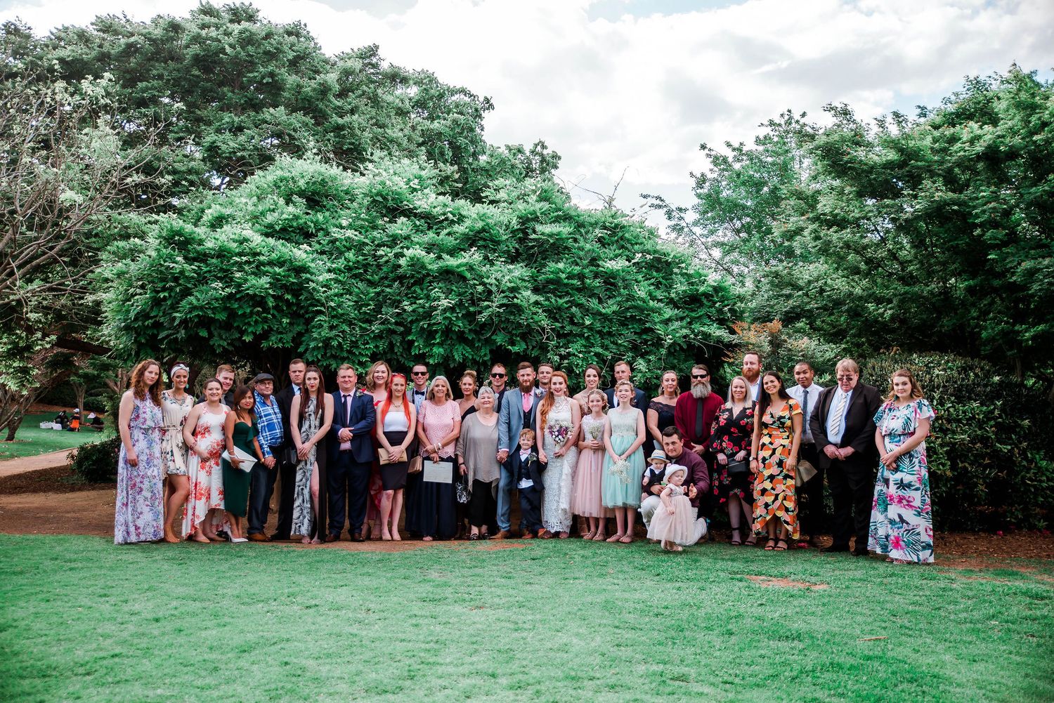 Large wedding party group photo on green lawn with trees in background.