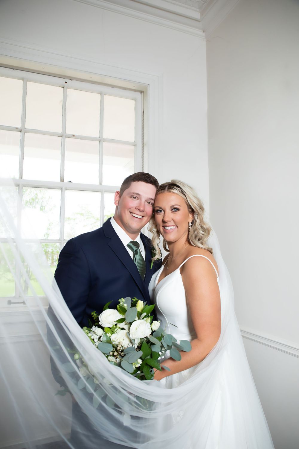 Elegant couple poses with white floral bouquet by window on their wedding day.