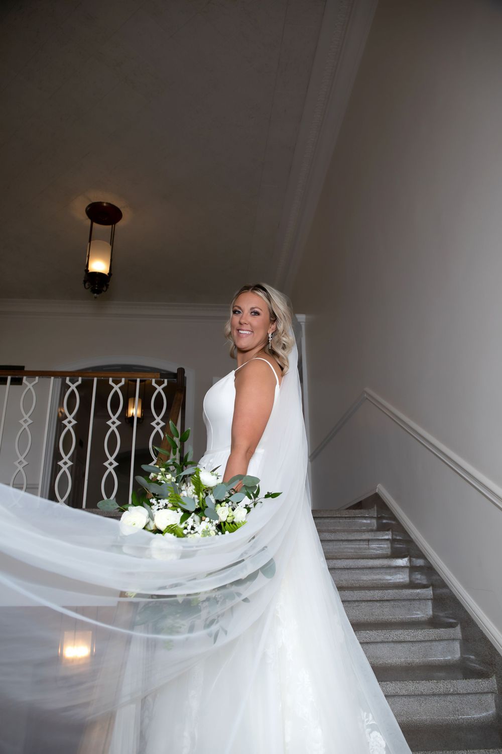 Bride with flowing veil and white bouquet poses on elegant staircase.
