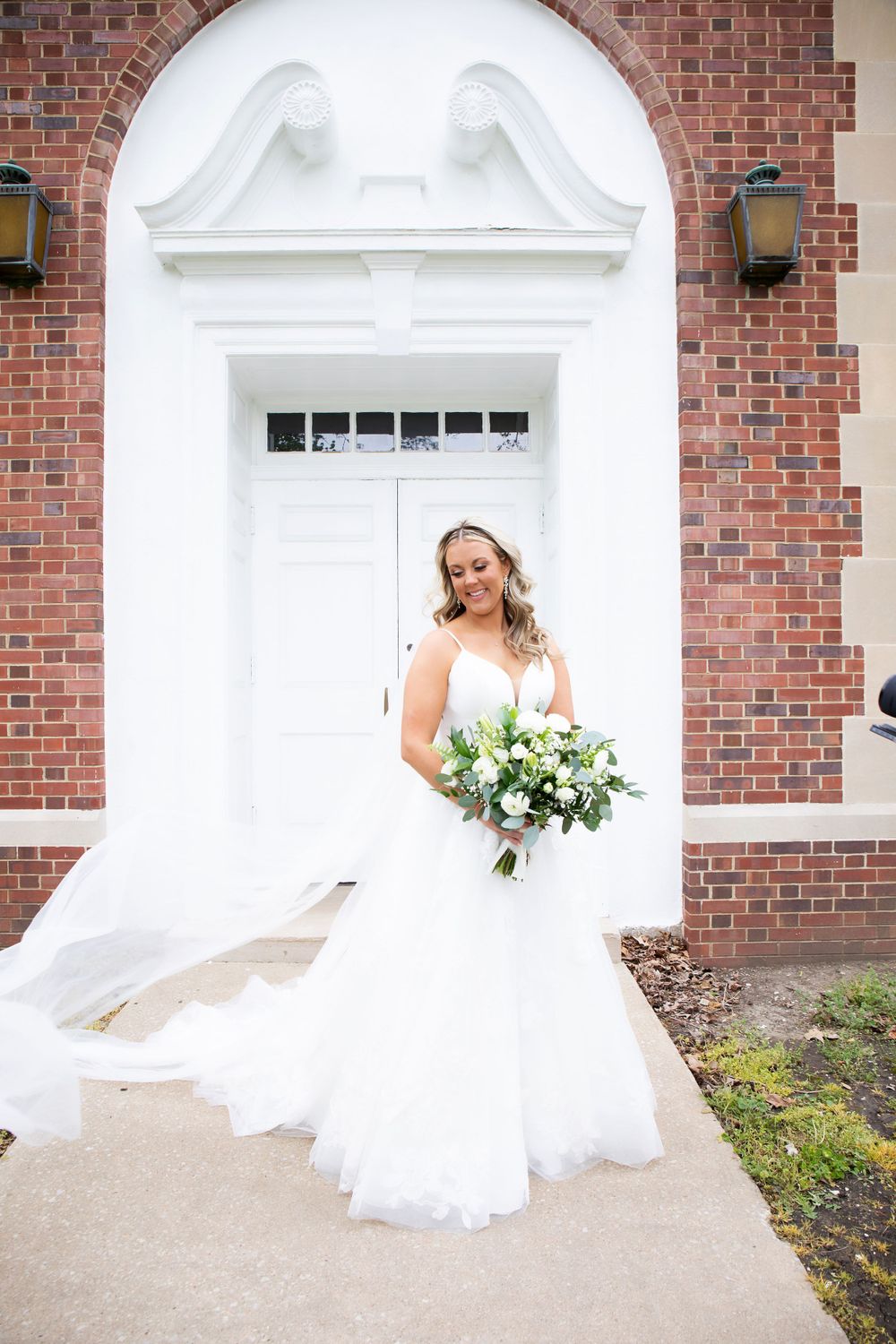 Bride with white bouquet poses in front of elegant arched church doorway.