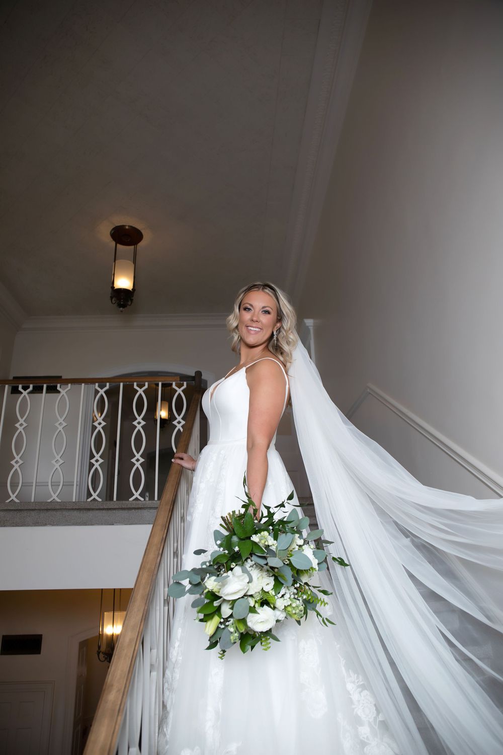 Bride in stunning white dress holds bouquet on staircase with flowing veil.