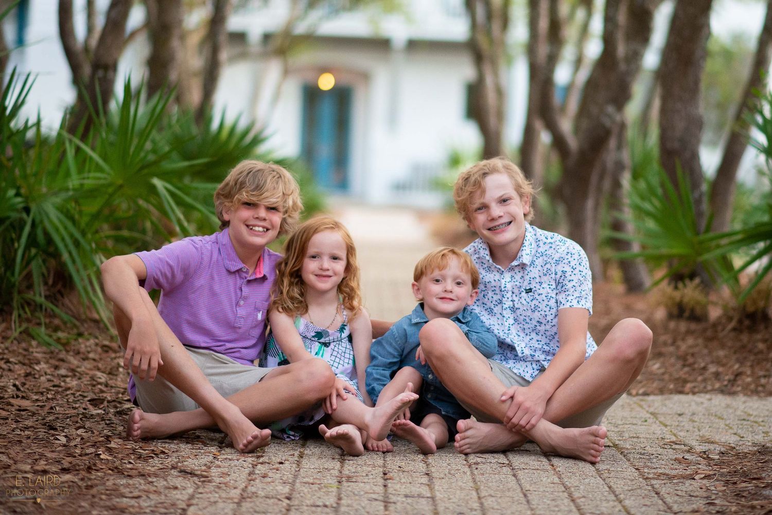 Children sitting together on a brick pathway surrounded by palm trees.