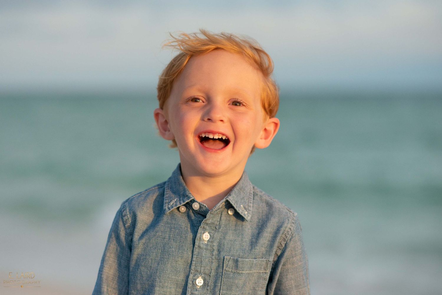 Child in denim shirt smiling with ocean in background.