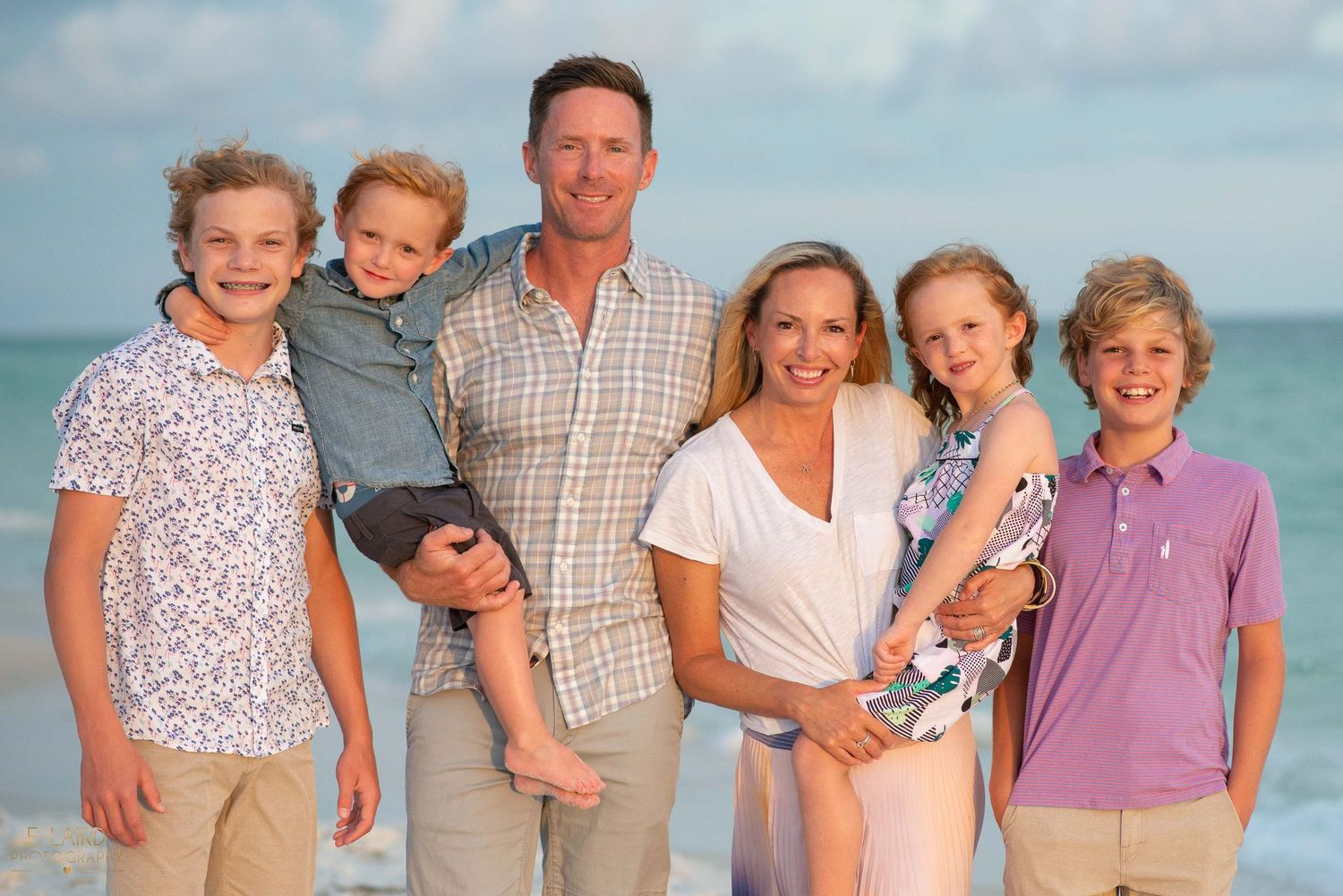 A family of six poses together on the beach at sunset wearing coordinated casual clothing.