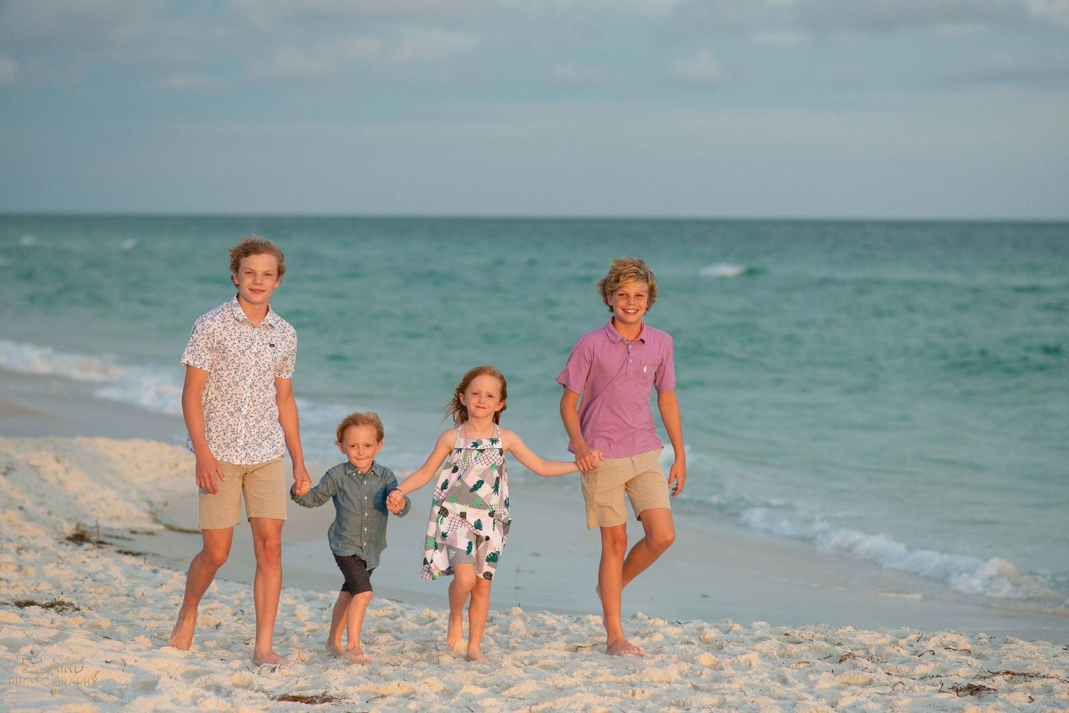 A family walks together along a scenic beach with turquoise waters and sand in casual summer attire.