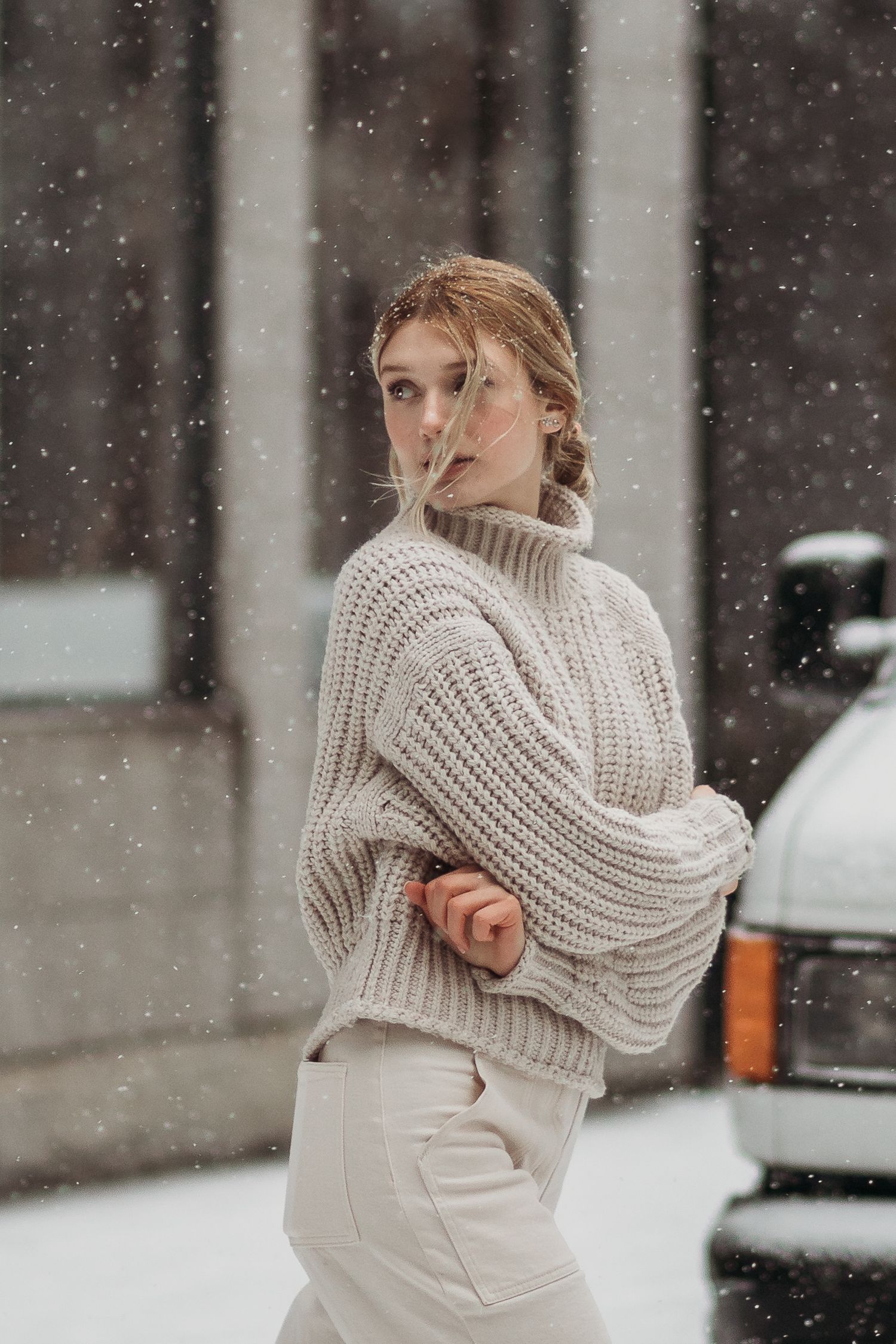 Side profile of someone in cream turtleneck sweater posing outdoors during snowfall.