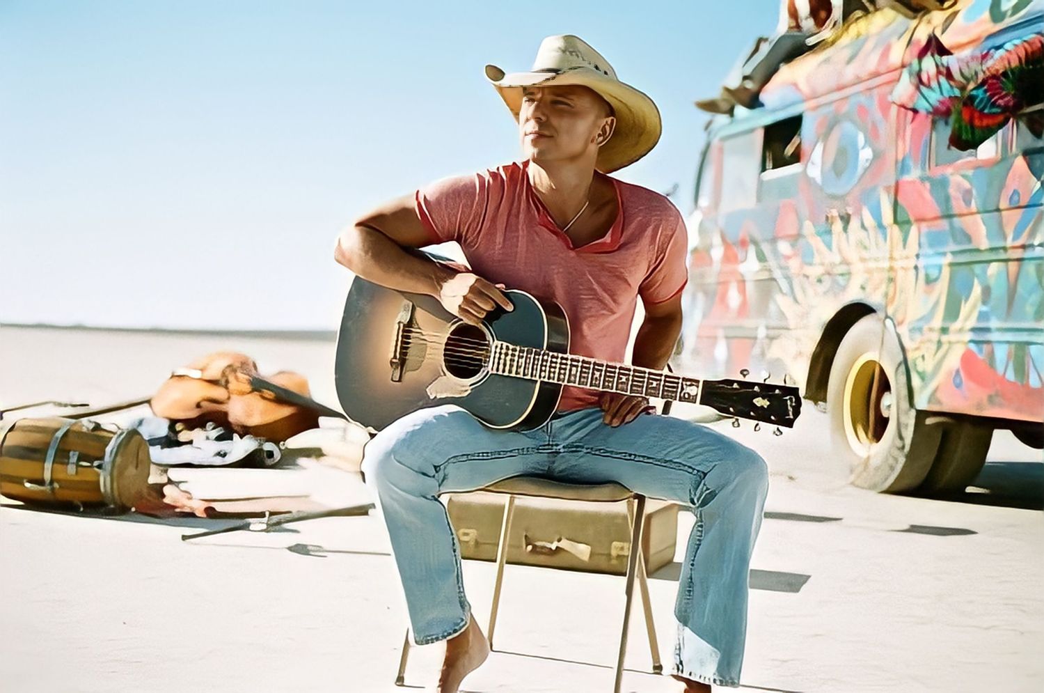 Musician in straw hat playing acoustic guitar while seated outdoors near colorful vehicle.