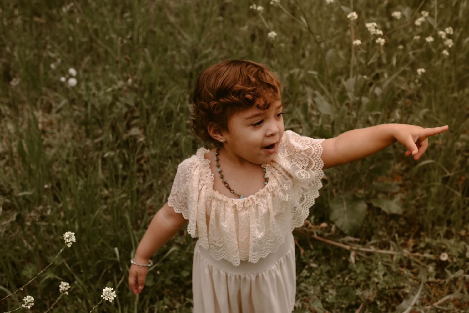 Child in white lace dress points while playing in a meadow of wildflowers.