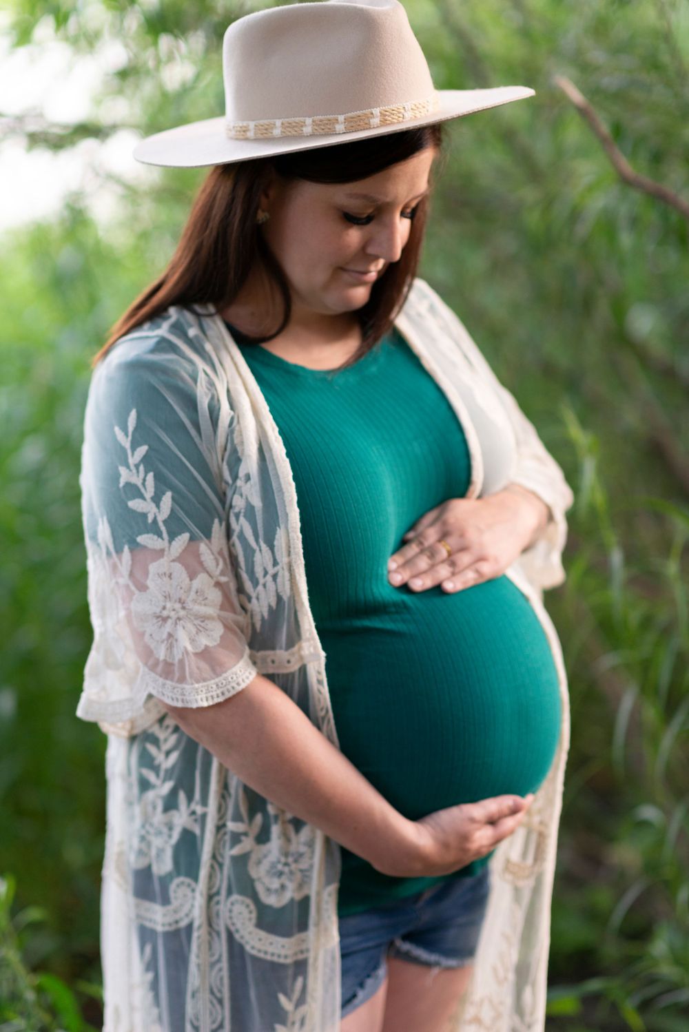 Maternity portrait featuring teal dress and lace overlay with wide-brimmed hat in garden setting.